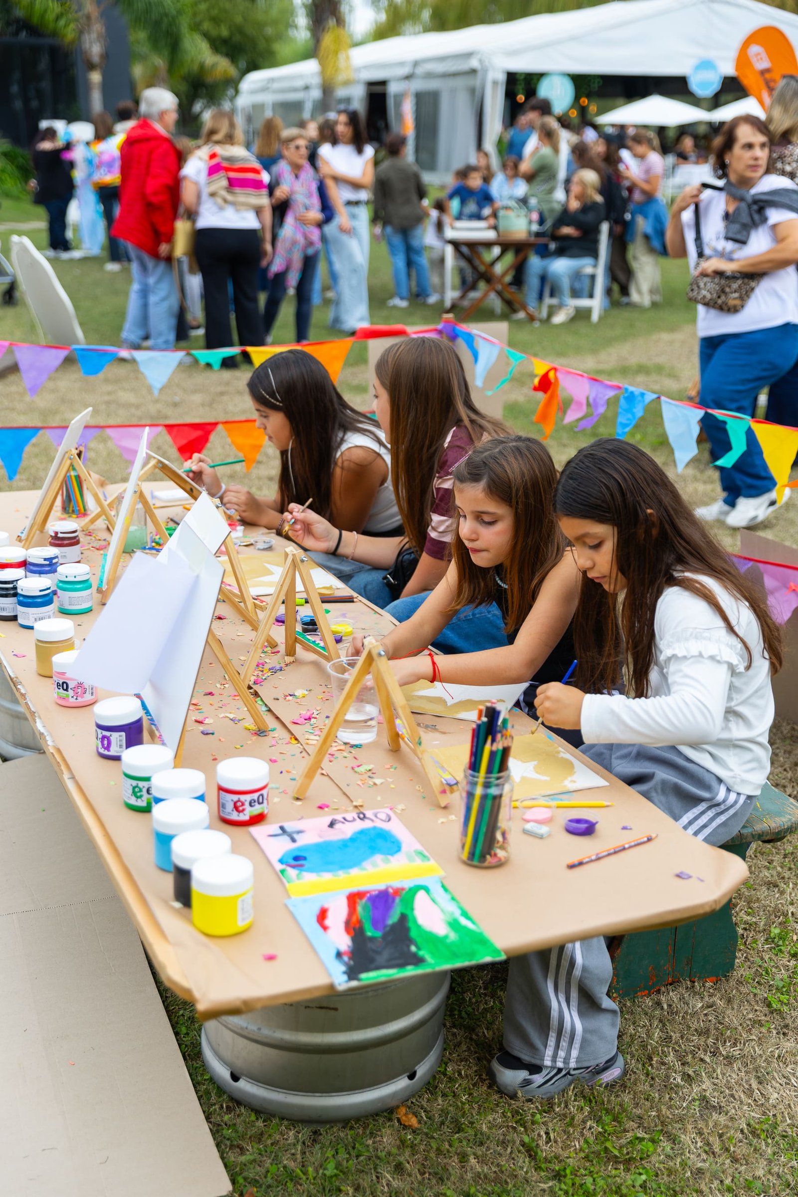 Chicas pintando en una mesa larga del predio, con la guirnalda de banderines del festival de fondo
