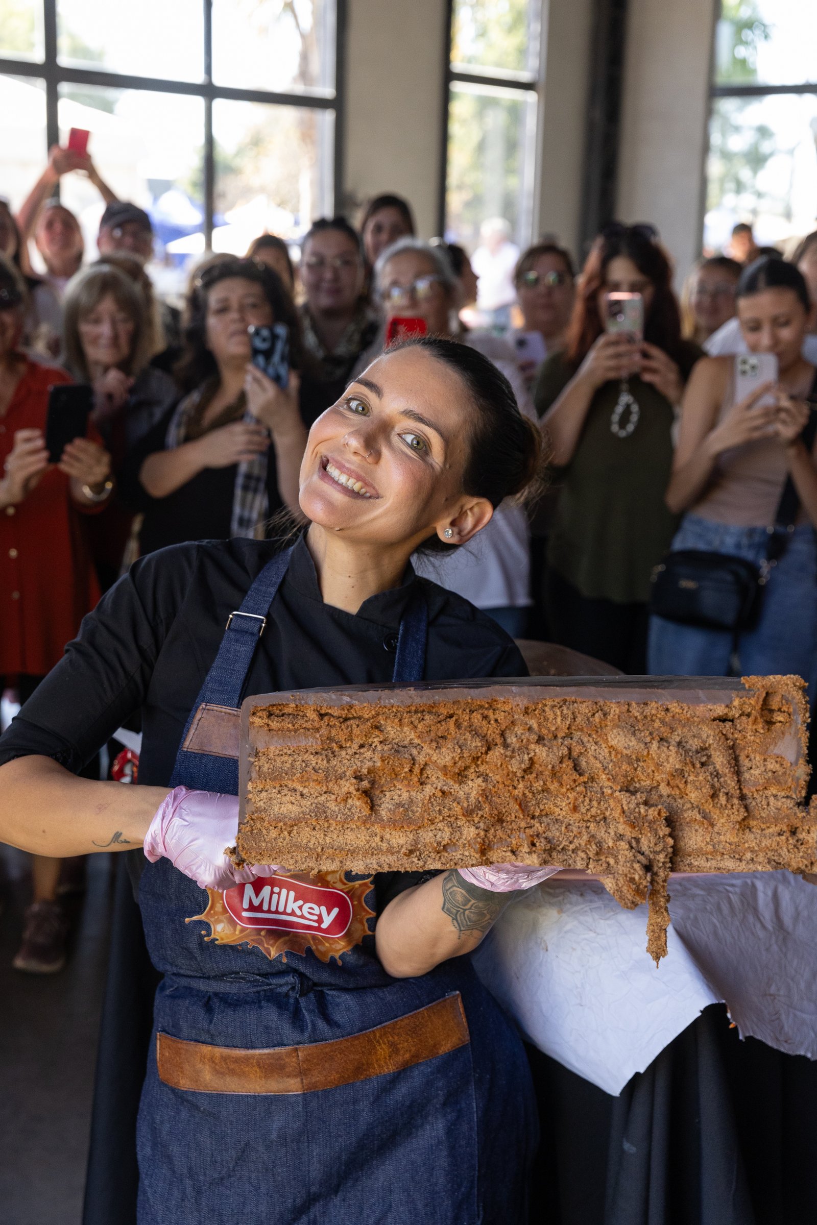 Chef sonriendo mientras muestra una torta enorme frente al público que la fotografía