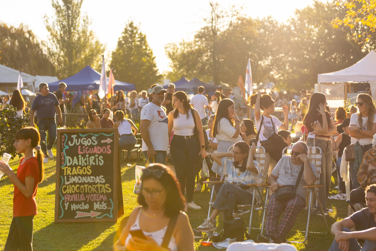 Multitud caminando entre los puestos del festival al atardecer junto al cartel de jugos