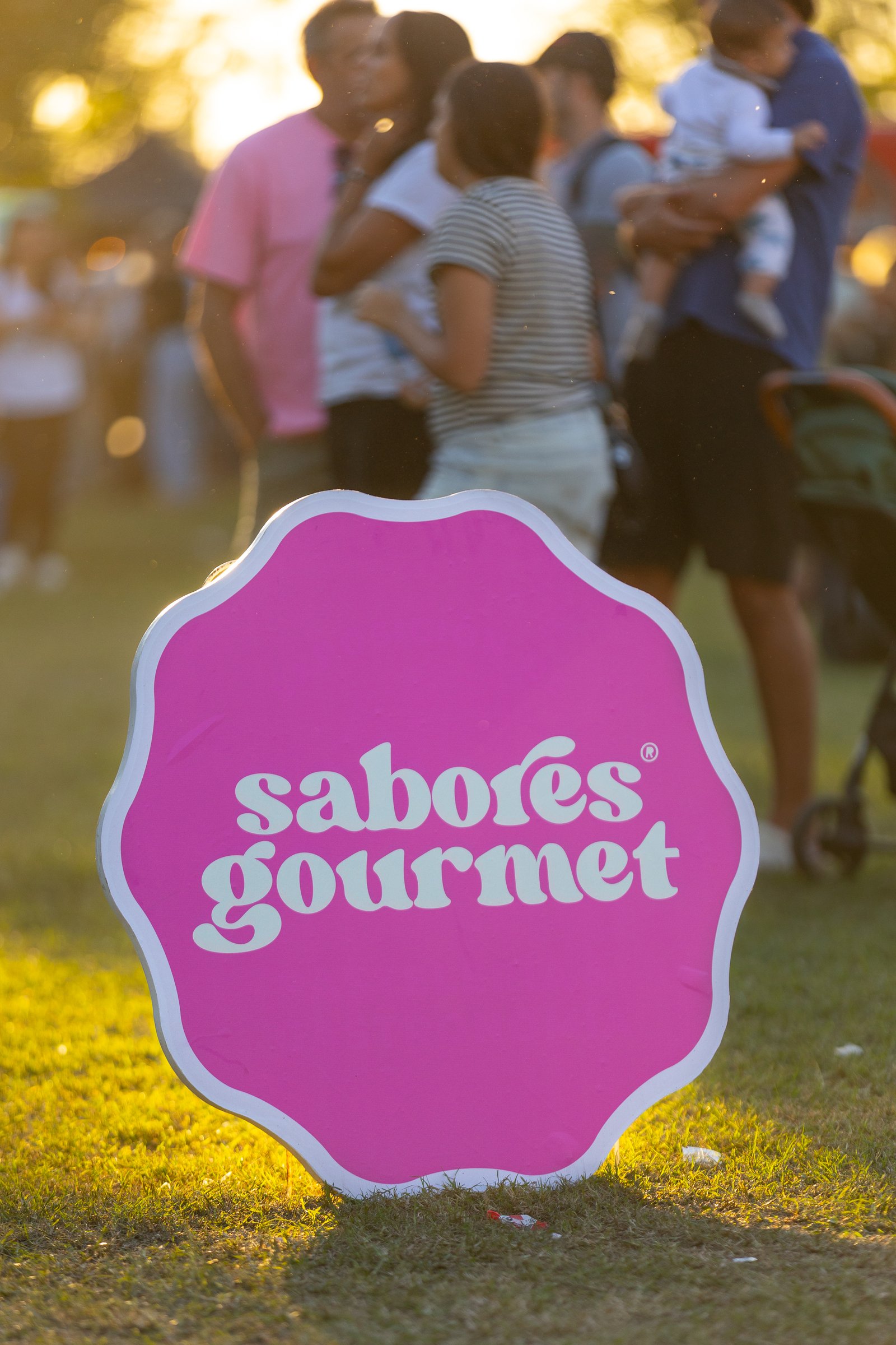 Cartel con el logo de Sabores Gourmet clavado en el pasto con gente de fondo